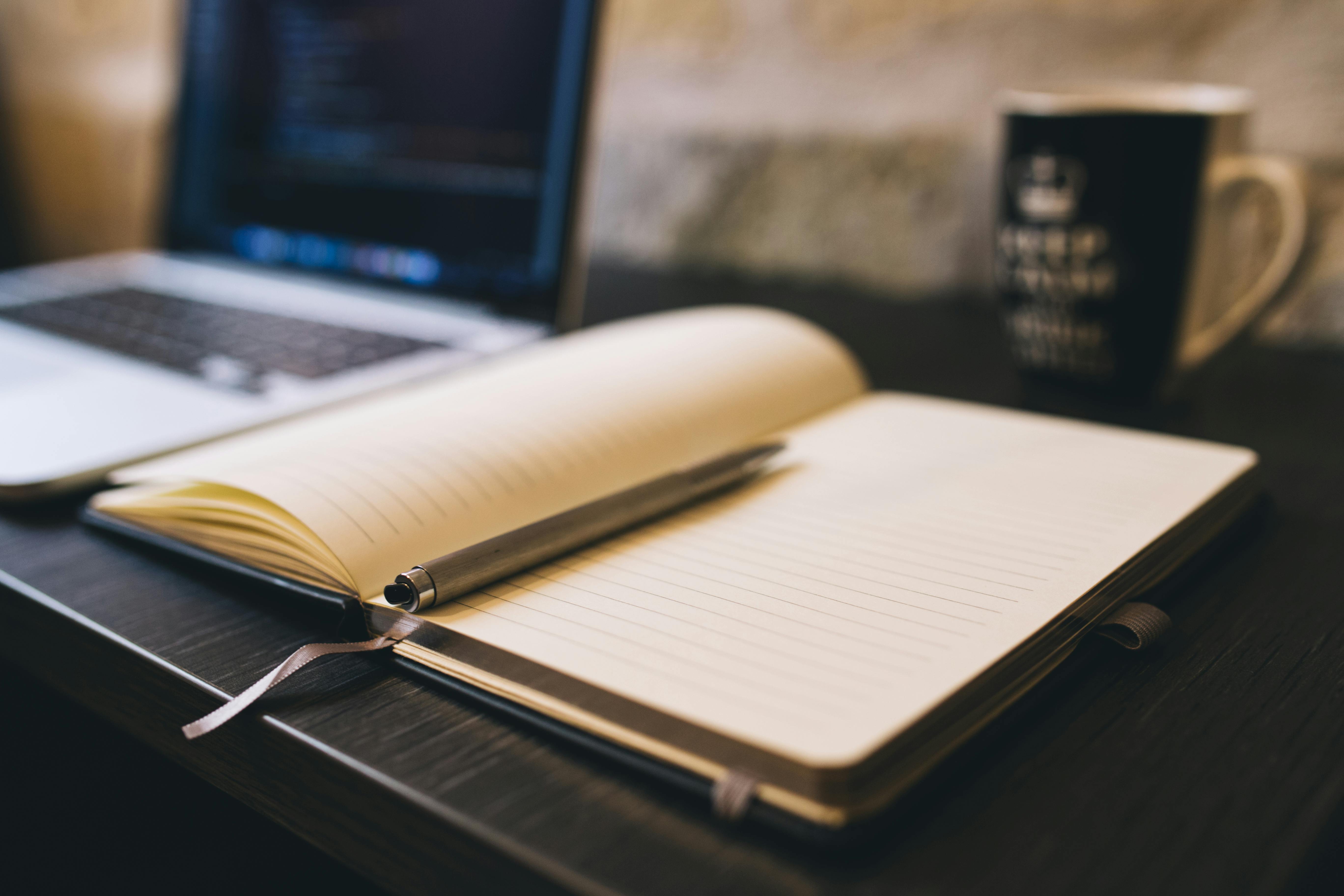 An open notebook with a pen, beside a laptop and coffee mug on a wooden desk